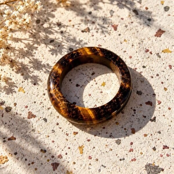Bague MELLIA anneau en œil de tigre poli, reflets miel et brun, posée sur pierre beige avec fleurs séchées
