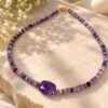 Collier en perles d'améthyste heishi lilas à violet, grande pierre centrale facettée et fermoir doré, posé sur pierre claire avec fleurs