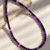 01-collier-perles-fines-amethyste Collier en perles fines d'améthyste violettes, fermoir et chaînette d'extension plaqué or, posé sur pierre claire avec ombres douces