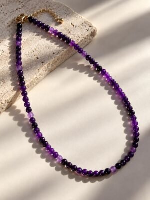 01-collier-perles-fines-amethyste Collier en perles fines d'améthyste violettes, fermoir et chaînette d'extension plaqué or, posé sur pierre claire avec ombres douces