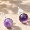 Puces d'oreilles en boules d'améthyste violette de 8 mm, tiges argentées et poussettes silicone, posées sur pierre beige