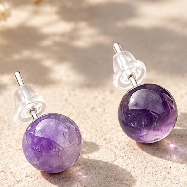 Puces d'oreilles en boules d'améthyste violette de 8 mm, tiges argentées et poussettes silicone, posées sur pierre beige