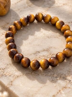 02-bracelet-mellia-oeil-de-tigre Bracelet MELLIA en perles d’œil de tigre posé sur une dalle de marbre clair, à côté d’une pierre brute