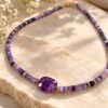 Collier perles améthyste INTUITION posé à plat sur dalle claire, pierre centrale facettée et perles dorées