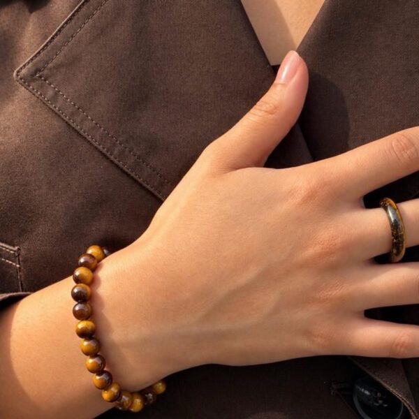 Bracelet MELLIA œil de tigre au poignet, main posée sur une chemise marron avec bague assortie