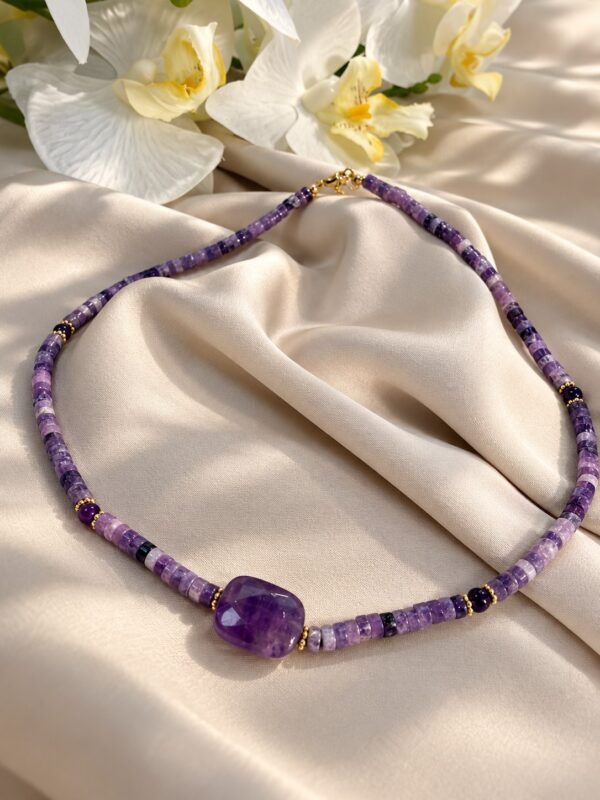 Collier Collection INTUITION en perles d'améthyste, pierre centrale facettée et détails dorés, posé sur satin avec orchidées