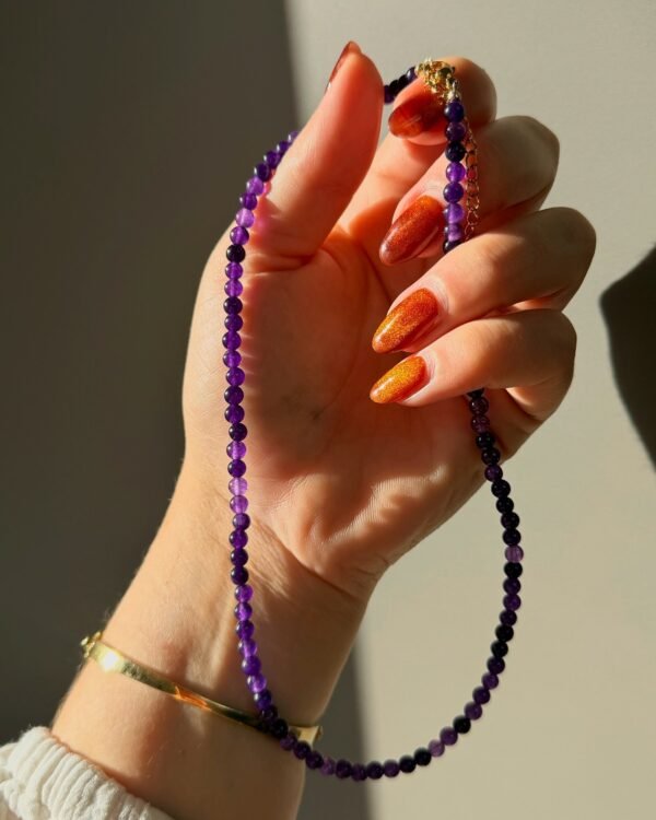 08-collier-perles-fines-amethyste Main tenant le collier perles fines améthyste, fermoir et chaînette de rallonge dorés, lumière naturelle