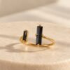 Bague NÉRA ouverte, deux pierres rectangulaires en obsidienne noire sur anneau en cuivre couleur dorée, posée sur pierre claire – vue 1