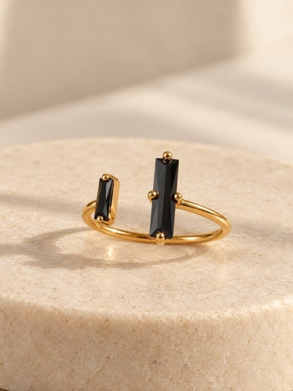 Bague NÉRA ouverte, deux pierres rectangulaires en obsidienne noire sur anneau en cuivre couleur dorée, posée sur pierre claire – vue 1