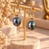 Boucles d’oreilles ÉPURE, grosses perles d’eau douce baroques noires aux reflets bleutés sur créoles finition ton or, présentées sur un support – vue 1