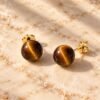 Boucles d’oreilles puces œil de tigre MELLIA Boucles d’oreilles puces MELLIA en œil de tigre naturel à bouchon, posées sur marbre clair – vue 1